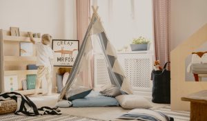 Real photo of a scandi playroom interior with a tent and pillows. Boy putting a toy on a shelf