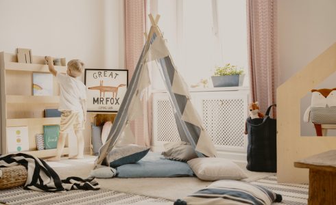 Real photo of a scandi playroom interior with a tent and pillows. Boy putting a toy on a shelf